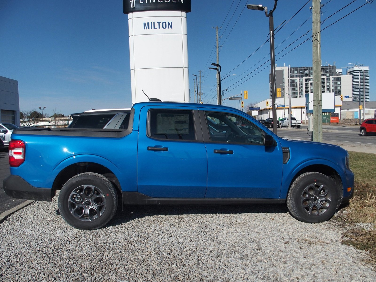 Side profile of a bright blue Ford Maverick pickup truck parked on gravel at a car dealership.